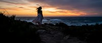 Woman standing on rocky coastal cliff at sunset, silhouetted against golden and purple sky, ocean waves below, contemplative travel and nature scene.
