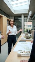 Professional middle-aged Caucasian female architect presenting urban development project in modern office with skylight and architectural models on desk.