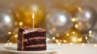 Chocolate birthday cake slice with lit candle on white plate surrounded by golden and silver balloons with warm bokeh lights creating festive atmosphere.