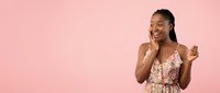 Young African American woman in floral dress smiling and touching face against pink background, expressing joy and confidence for lifestyle content.