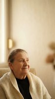 Senior woman with light skin tone and gray hair wearing cream cardigan and black turtleneck, sitting by window in warm natural light, expressing contentment and peaceful aging in home interior setting.