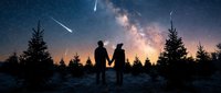 Couple holding hands gazing at starry night sky with milky way and meteor shower above snowy forest landscape at dusk.