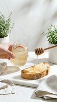 Person with light skin tone holding jar of honey with wooden dipper over toasted bread slice on white plate, fresh herbs in pots, natural sunlight, breakfast or food styling setup.