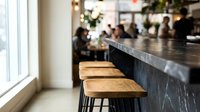 Young adult female with medium skin tone sitting at modern bar counter with wooden seat and black metal legs in contemporary café setting with blurred background patrons and warm lighting for hospitality and lifestyle use.