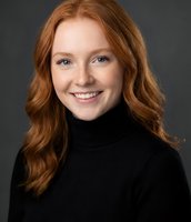Young Caucasian woman with curly red hair smiling warmly in black turtleneck sweater against gray background for professional headshot and portrait photography.