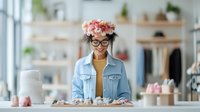Cheerful female entrepreneur with flower crown and glasses in light blue denim jacket standing behind counter in boutique shop with handmade goods.