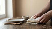 Male hands kneading dough on wooden table with ceramic bowls in bright kitchen interior for homemade bread baking and cooking preparation.