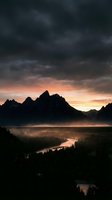 Dramatic mountain landscape at sunrise with silhouetted peaks, golden sky breaking through dark storm clouds, and misty river valley below for nature and travel projects.