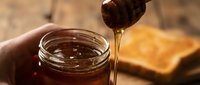 Honey dripping from wooden dipper into glass jar with toast in background, sweet food ingredient for cooking and baking projects.