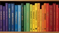 Colorful rainbow arrangement of books on wooden shelf creating spectrum from purple to red spines for education and learning concepts.