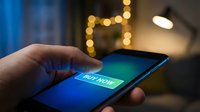 Hand holding smartphone with glowing buy now button on screen, online shopping concept in cozy home setting with bokeh lights.