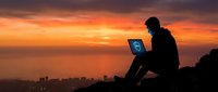 Silhouetted male professional working on laptop against dramatic golden sunset sky overlooking city skyline, representing remote work, entrepreneurship, and digital connectivity.