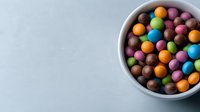 Colorful chocolate candy pieces in white ceramic bowl on light background, overhead view of round multicolored sweets for confectionery and dessert designs.