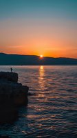 Silhouette of person standing on coastal cliff edge watching dramatic orange sunset over mountain range and calm sea with golden light reflection on water surface.