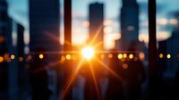 Blurred urban cityscape at sunset with bright sun flare through office building windows creating dramatic bokeh lights and warm golden hour atmosphere.
