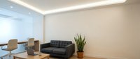 Modern office interior with minimalist design featuring black leather sofa, wooden desk, potted plant, and glass partition wall with ambient ceiling lighting.