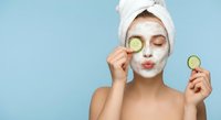 Young woman with white towel on head applying facial mask holding cucumber slices for skincare routine on blue background for beauty and wellness content.