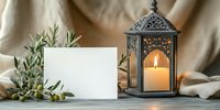 Decorative lantern with glowing candle and blank white card on table with olive branches and beige fabric backdrop, creating cozy atmosphere.