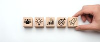 Hand placing wooden cube with rocket icon completing business strategy sequence with team, idea, growth, target symbols on white background.