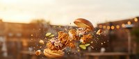 Freshly baked burger halves with sesame seeds exploding mid-air against warm sunset backdrop with bokeh lights, perfect for food advertising and restaurant marketing campaigns.