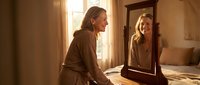 Young blonde woman in brown sweater smiling at her reflection in wooden framed mirror in warm sunlit bedroom for self-care and confidence concepts.