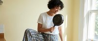 Young mixed-race woman with curly hair sitting on wooden floor holding black frying pan in bright kitchen interior with natural window light.