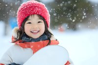 Cheerful Asian child wearing pink winter hat and orange jacket holds snowboard while enjoying snowy weather, expressing pure joy during winter sports activity.