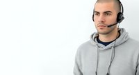 Young man with dark hair and facial hair wearing gray hoodie and headset with microphone, isolated on white background for customer service, tech support, or call center applications.