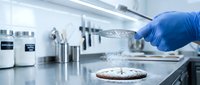 Scientist in blue glove pouring liquid onto petri dish in laboratory setting with equipment and instruments on bench.