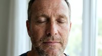 Middle-aged Caucasian man with closed eyes and peaceful expression practicing mindfulness meditation indoors by window with natural light for wellness and relaxation.