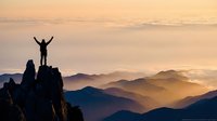 Male hiker celebrating success on mountain peak at sunrise with arms raised, overlooking misty valley layers and distant mountain ranges in golden light.
