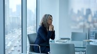 Professional blonde businesswoman in navy suit contemplating by office window with city skyline view, corporate leadership and strategic planning concept.
