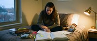 Young woman studying with books and papers on bed in cozy bedroom with warm lamp light during evening hours for academic research and learning.
