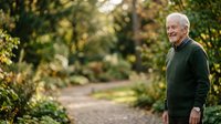Senior man with white hair and light skin tone, wearing dark green sweater, smiling while standing on garden pathway surrounded by lush green foliage and trees during daytime.