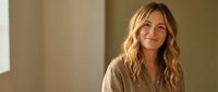Happy blonde woman with wavy hair smiling warmly in casual beige shirt against soft neutral background for lifestyle and wellness content.