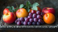 Fresh seasonal fruits arranged on ornate vintage silver tray against dark background. Red apples, oranges and purple grapes with green leaves create classical still life composition.