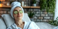 Young Asian woman relaxing at spa with facial mask and cucumber slices on eyes, wearing white bathrobe and towel, enjoying skincare treatment at wellness center.