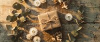 Rustic gift box wrapped in kraft paper with twine bow surrounded by dried eucalyptus leaves and white flowers on weathered wooden background for holidays.