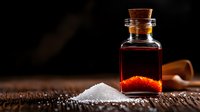 Small glass bottle with red liquid and cork stopper next to pile of sea salt crystals on rustic wooden surface against dark background, dramatic lighting.