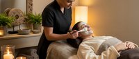 Professional spa therapist performing relaxing facial treatment on client in luxury wellness center with candles and plants creating serene atmosphere.