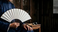 Person in dark blue traditional garment holding open white and black folded fan, displaying craftsmanship and cultural heritage for editorial and lifestyle content.