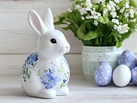 White ceramic bunny figurine decorated with blue floral pattern sits on wooden surface near Easter eggs and spring flowers in vintage pot. Seasonal home decor arrangement.