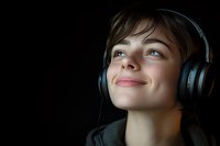 Young Caucasian woman with short brown hair wearing wireless headphones smiles while looking up, illuminated against dark background, expressing joy and relaxation.