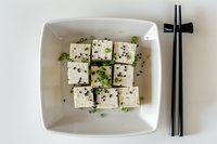 Fresh tofu cubes garnished with green onions and sesame seeds served in white ceramic bowl with black chopsticks for healthy Asian cuisine recipes.