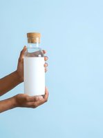 Hand holding glass bottle with cork stopper and white label against light blue background for product mockup and branding.