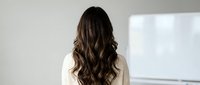 Rear view of professional businesswoman with long wavy brown hair in white blazer standing in modern office environment for corporate presentations.