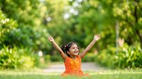 Young girl with dark hair and medium skin tone, wearing orange dress, arms raised in celebration outdoors among green trees and grass on sunny day, expressing joy and freedom.