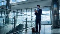 Professional businessman in suit using smartphone while waiting with luggage in modern airport terminal or office building with large windows and city view.