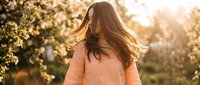 Young woman with long brown hair in motion wearing coral sweater during golden hour sunset in blooming spring garden with warm natural lighting.
