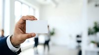 Businessman holding blank white business card in modern office environment with colleagues and plants in background.